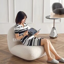 Một phụ nữ trẻ đang ngồi thư giãn trên ghế GTE38 màu trắng kem, đọc sách. Ghế có kiểu dáng hiện đại, bọc vải giả len mềm m…