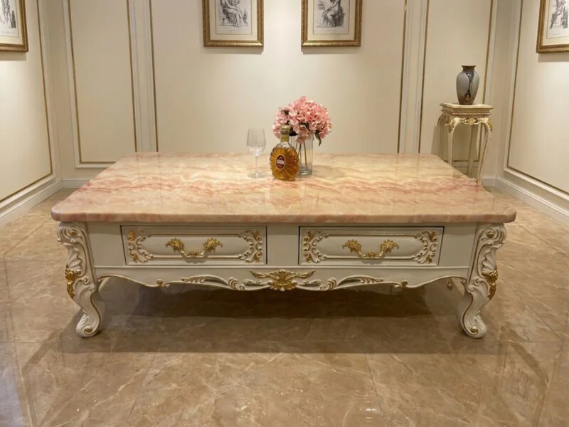 Bàn trà BELLAROSE tân cổ điển đặt giữa phòng khách sang trọng với sàn đá cẩm thạch, tường trắng và tranh treo tường. Mặt b…
