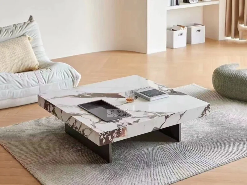 Bàn trà vuông mặt đá nhập khẩu GK04-1M được chụp từ góc hơi chếch phía trước, đặt trên thảm trải sàn màu xám nhạt trong mộ…