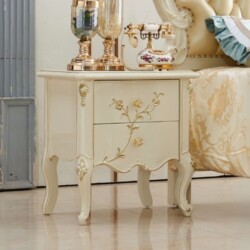 Tủ đầu giường TAD02 màu trắng kem với các chi tiết chạm khắc vàng đồng, đặt cạnh giường ngủ bọc nệm màu kem. Trên mặt tủ c…