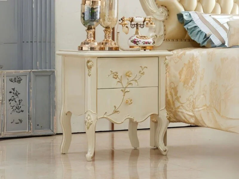Tủ đầu giường TAD02 màu trắng kem với các chi tiết chạm khắc vàng đồng, đặt cạnh giường ngủ bọc nệm màu kem. Trên mặt tủ c…