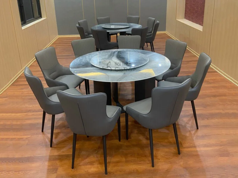 Bộ bàn ăn tròn hiện đại BA928-135 chụp chính diện, với mặt bàn đá vân xám đen có mâm xoay. Chân bàn trụ màu đen. Xung quan…