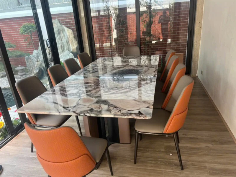 Bàn ăn mặt đá tự nhiên BAQ55-24 được đặt trong không gian phòng ăn hiện đại, cạnh cửa kính lớn nhìn ra sân vườn. Mặt bàn h…