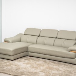 Sofa da góc phải màu be/ghi sáng, chụp từ góc thấp phía trước bên trái, đặt trên thảm lông màu kem trong không gian phòng…