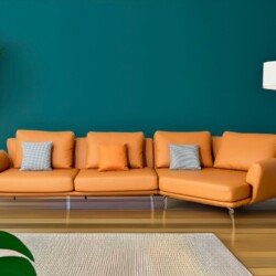 Bộ sofa da góc màu cam được chụp chính diện, đặt trên sàn gỗ sáng màu với thảm trải sàn họa tiết, nổi bật trên nền tường x…