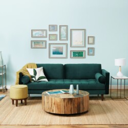 Sofa băng 3 chỗ bọc vải màu xanh lá cây đậm, đặt trong phòng khách ấm cúng với tường xanh nhạt, sàn gỗ và thảm họa tiết. G…