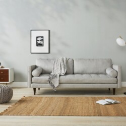 Sofa băng 3 chỗ bọc vải màu xám nhạt, đặt trong phòng khách tối giản với tường trắng, sàn gỗ và thảm cói. Góc chụp ngang…