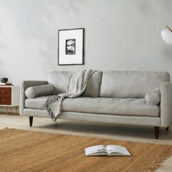 Sofa băng 3 chỗ bọc vải màu xám nhạt, đặt trong phòng khách hiện đại với tường trắng, sàn gỗ và thảm cói. Góc chụp ngang…