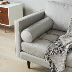 Cận cảnh tay vịn và gối ôm tròn của sofa bọc vải màu xám nhạt, đặt trong phòng khách hiện đại. Chi tiết đường may tinh tế…