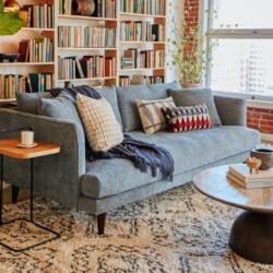 Sofa băng Lewis Extra SF-646 màu xám xanh, đặt trong phòng khách hiện đại với tường gạch, kệ sách lớn và bàn trà tròn, tạo…