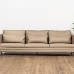 Ghế sofa băng ba chỗ BM001-113-C1050 màu nâu be với ba đệm tựa lưng rời, đặt trên sàn gỗ tối màu, phía sau là bức tường tr…