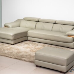Sofa da góc phải màu be/ghi sáng, chụp từ góc cao phía trước bên phải, đặt trên thảm lông màu kem và sàn gạch. Kèm theo mộ…