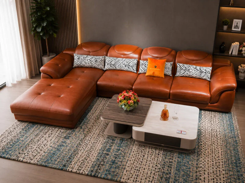 Góc nhìn từ trên xuống của bộ sofa góc chữ L bọc da màu nâu cam, nổi bật với các gối tựa họa tiết ngựa vằn và gối cam. Bàn…